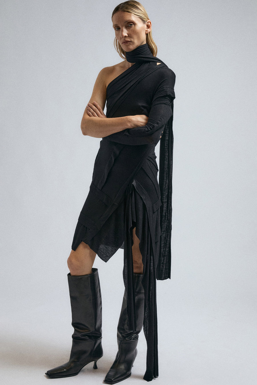 Woman wears JordanLuca black wrap dress, draped around the neck, cascading down the leg. 