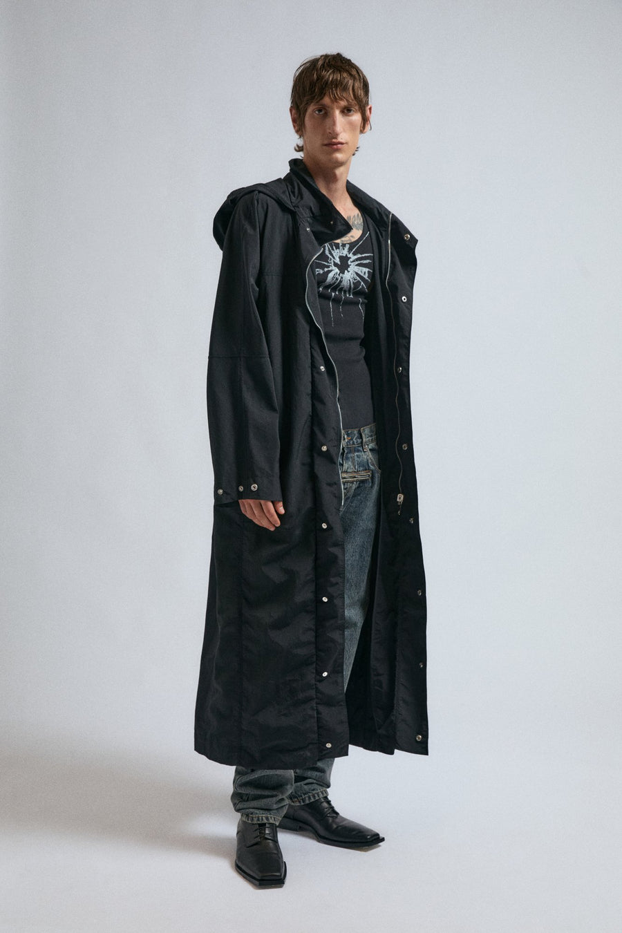 Man wears the black JordanLuca Megabit Duster, paired with the Shatter Tank Top, and the Marble Robin Jeans.